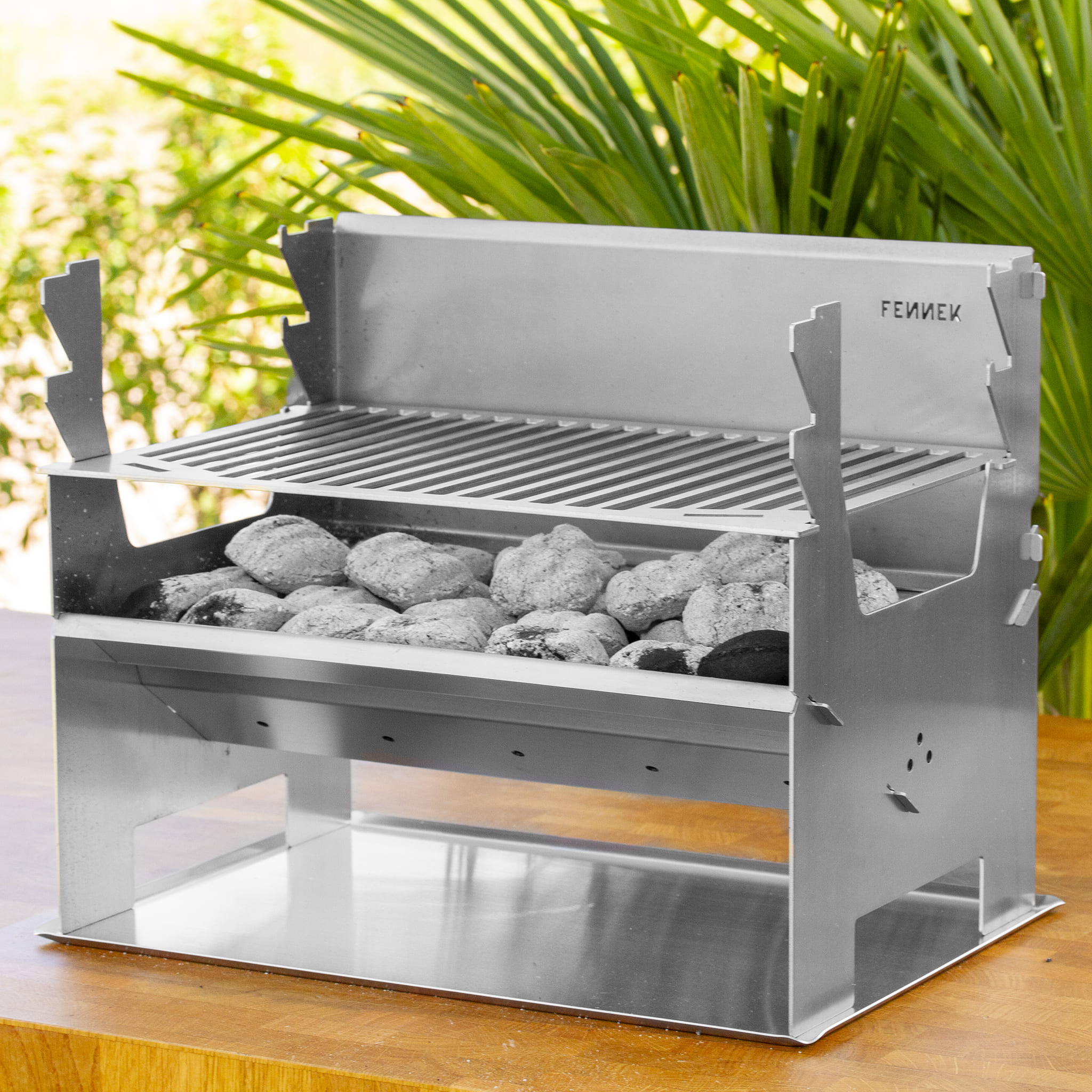 Tragbarer Edelstahl-Holzkohlegrill mit heißen Kohlen auf einem Holztisch vor grünen Blättern.