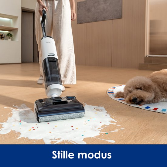 Een persoon maakt gemorste melk op een houten vloer schoon met een stofzuiger, terwijl een hond op een mat rust.
