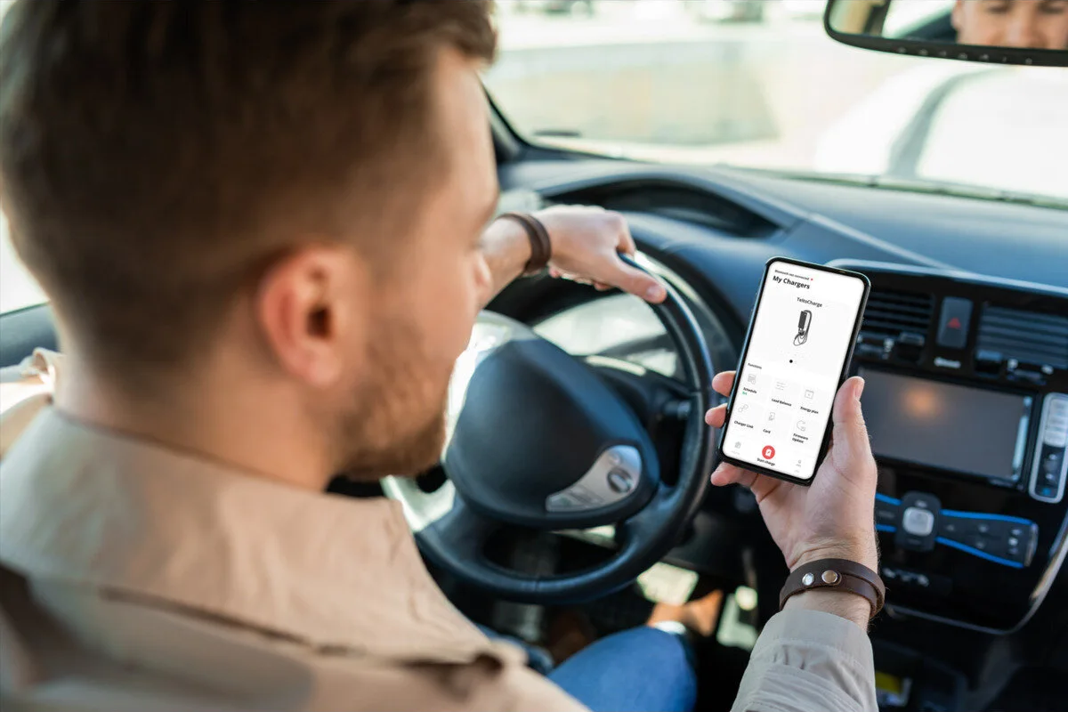 Bir arabanın direksiyonundaki adam, şarj uygulaması açık bir akıllı telefon tutuyor.