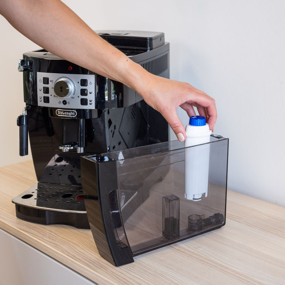 A hand places a water filter into the removable clear plastic water tank of a black DeLonghi espresso machine.