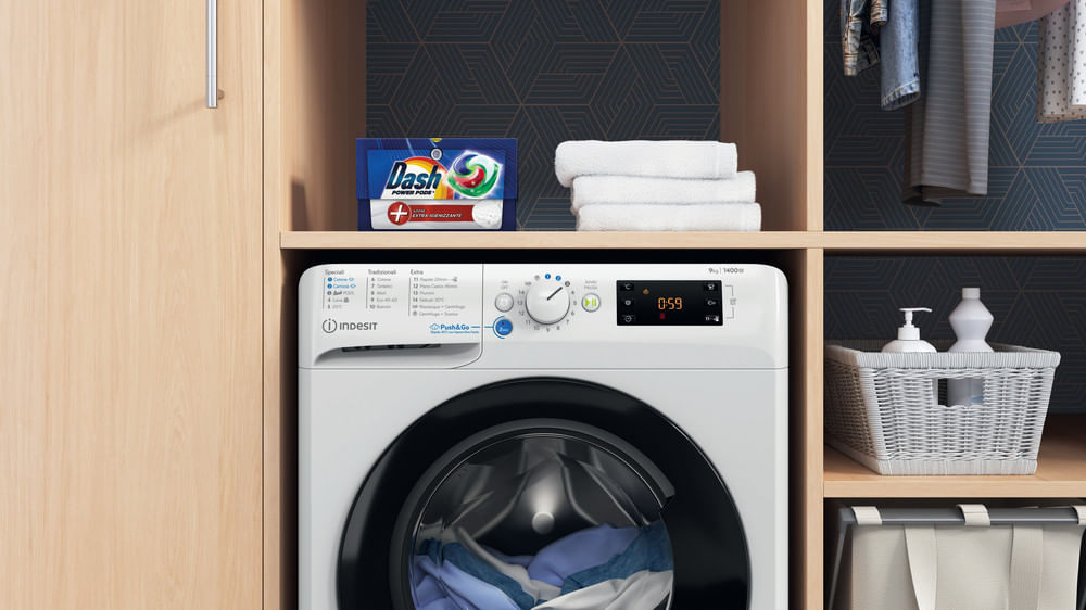 Una lavatrice bianca Indesit si trova in un mobile in legno con capsule di detersivo, asciugamani e un cestino.