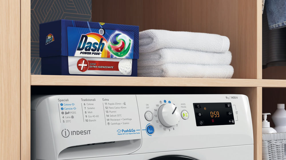 Una lavatrice Indesit bianca sotto un ripiano con una scatola di detersivo Dash blu e asciugamani bianchi piegati.