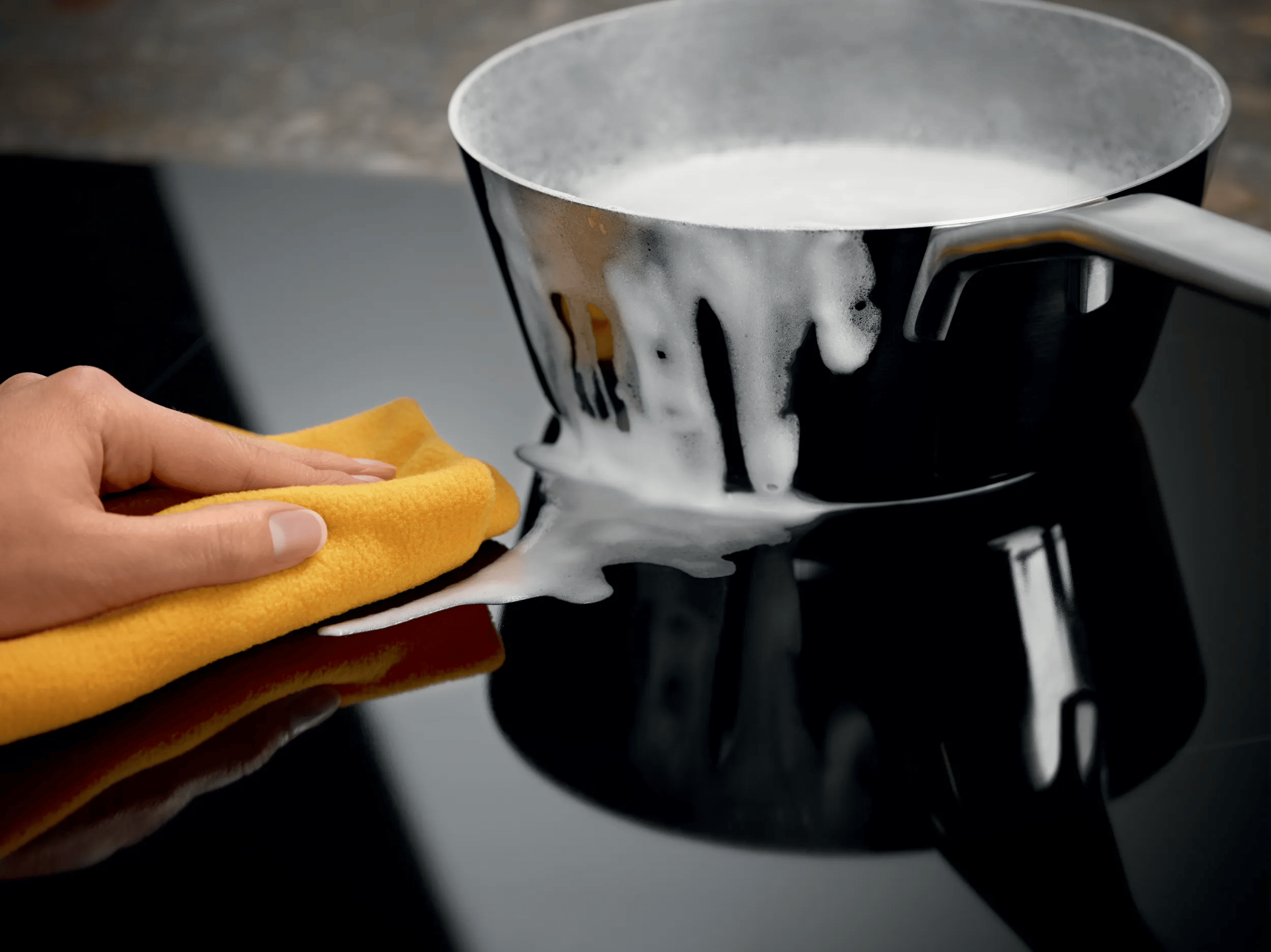Una mano usa un panno giallo per pulire il latte bollente versato su un piano cottura nero.
