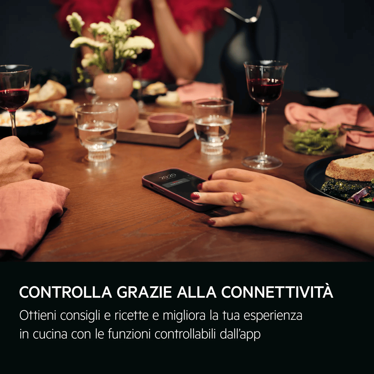 Persone a cena a un tavolo di legno con calici di vino, cibo, fiori e uno smartphone.