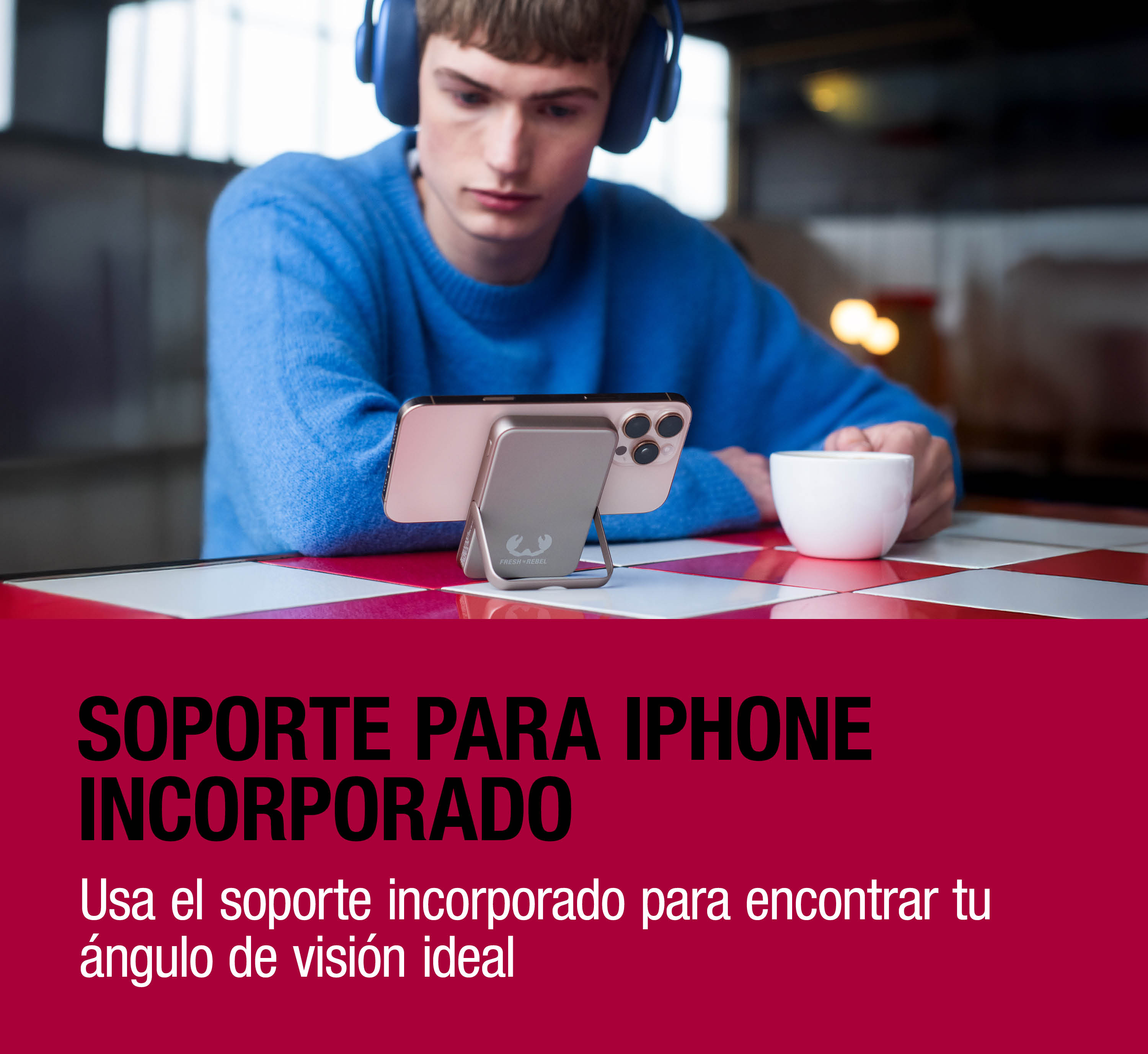 Un uomo con maglione blu e cuffie guarda uno smartphone su un supporto argentato, con una tazza accanto.