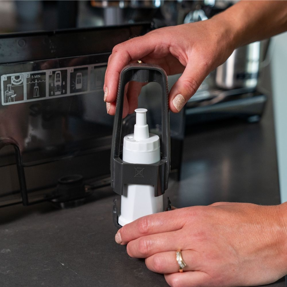 Hands place a white water filter into a dark grey holder on a dark surface, with a coffee machine in the background.