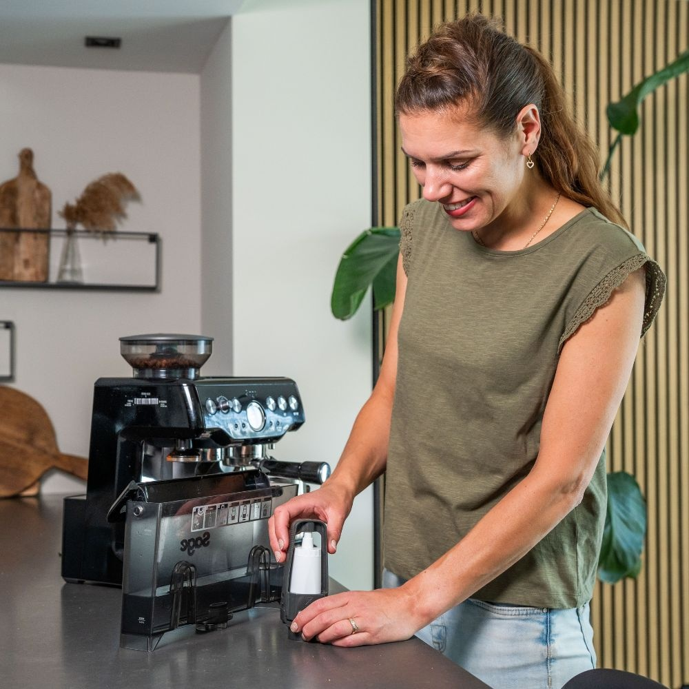 A woman in a green top holding a water filter in front of a coffee machine in a kitchen.