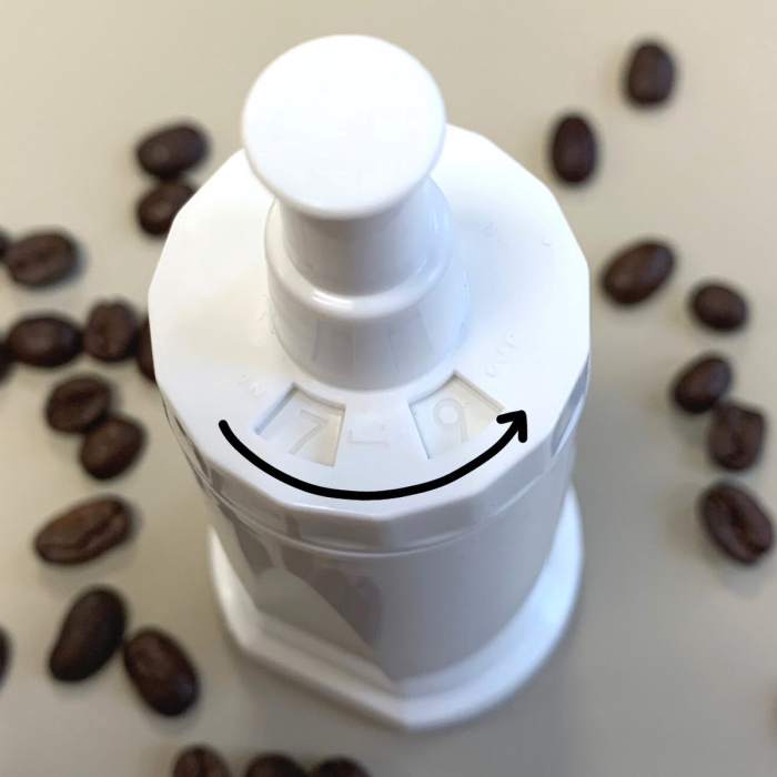 A white grinder part is shown with numbered settings and coffee beans scattered around it on a light surface.