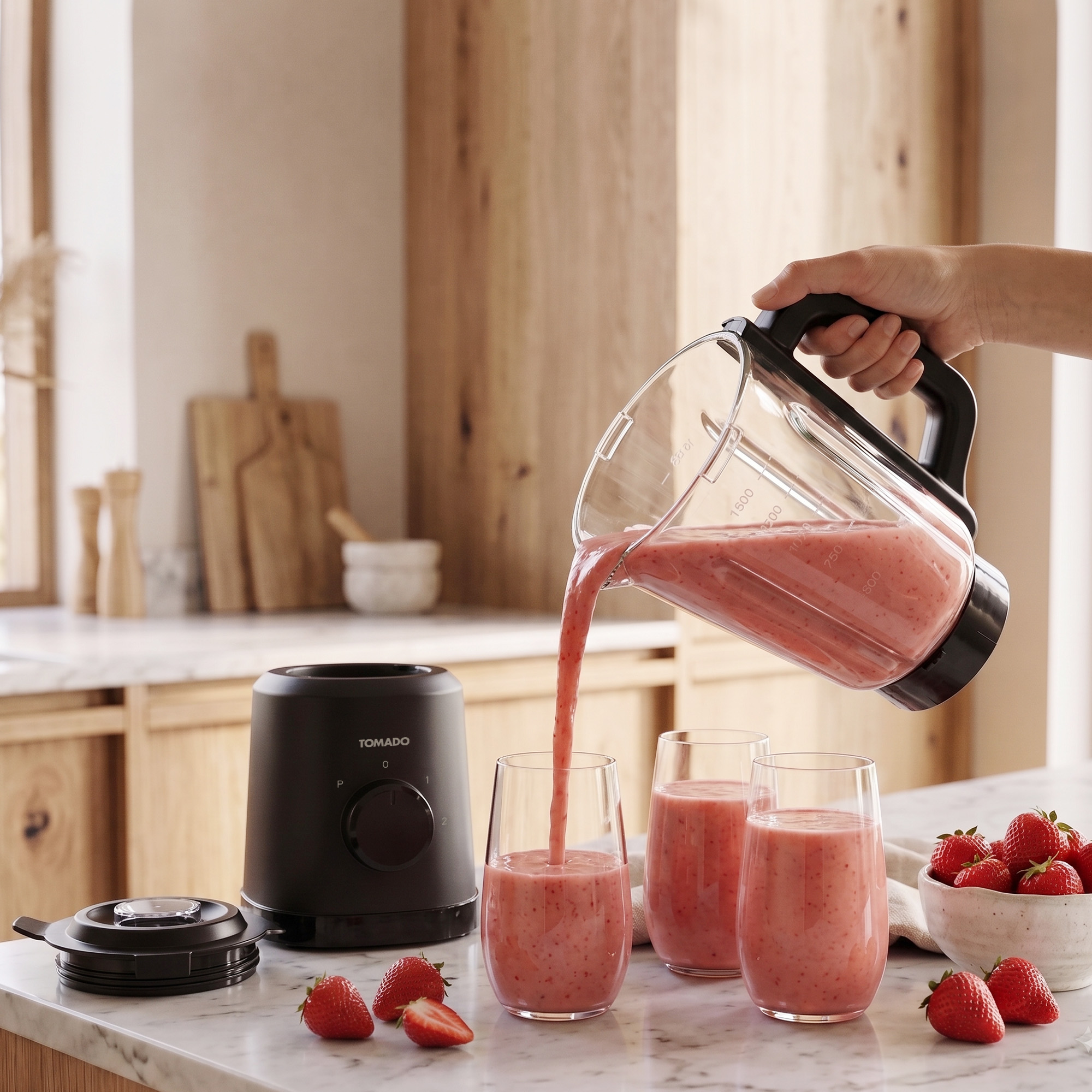 A hand pours a pink strawberry smoothie from a blender pitcher into a glass on a marble counter with fresh strawberries.