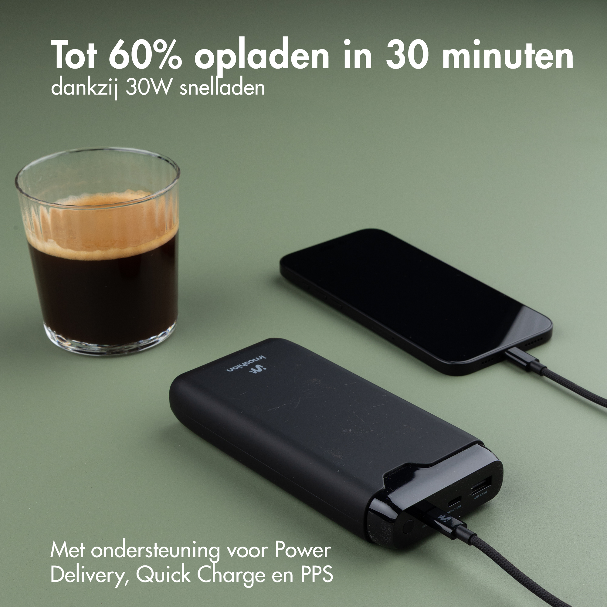 Eine schwarze Powerbank lädt ein Smartphone auf grüner Oberfläche neben einem Glas Kaffee mit niederländischem Text.