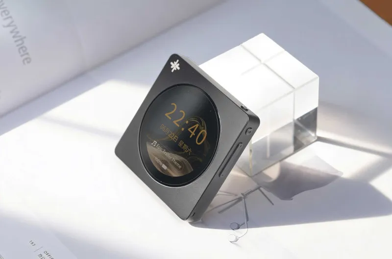 Un reproductor de música negro cuadrado con pantalla redonda y un cubo decorativo blanco sobre superficie blanca.