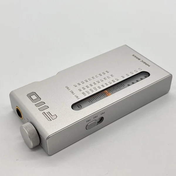 Un dispositivo de audio FiiO plateado con escala analógica, perilla y interruptor sobre fondo claro.