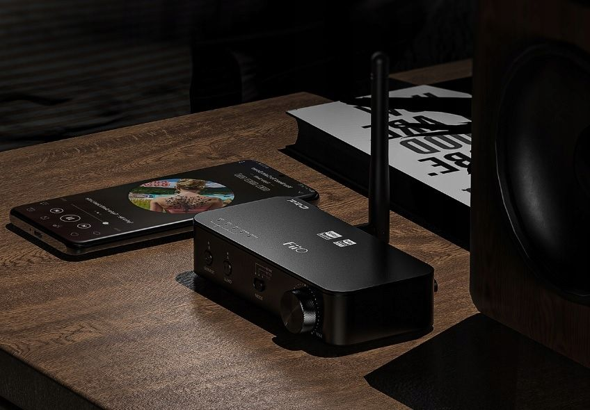 Un amplificador de audio FiiO con antena en una mesa de madera, con un smartphone y un altavoz cerca.