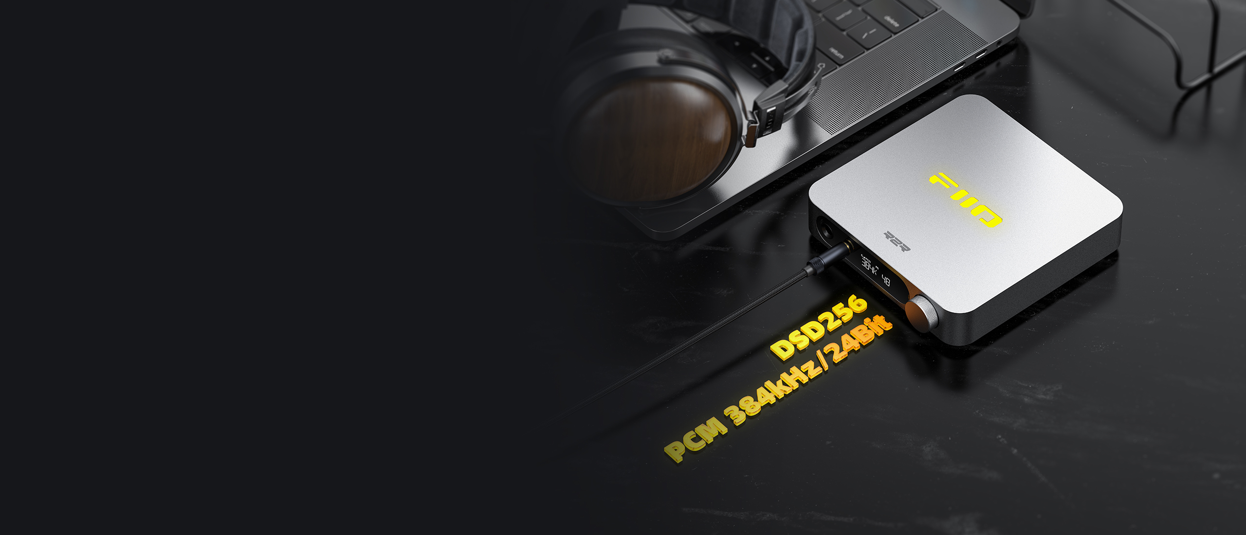 Srebrny odtwarzacz audio z żółtym logo na czarnym stole, obok laptopa i słuchawek.