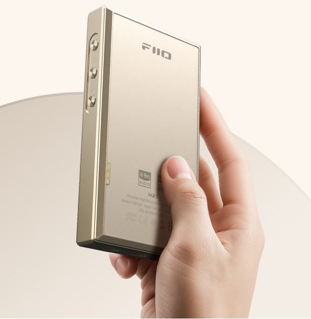 Una mano sostiene un dispositivo de audio portátil dorado con botones laterales y texto en la parte posterior.