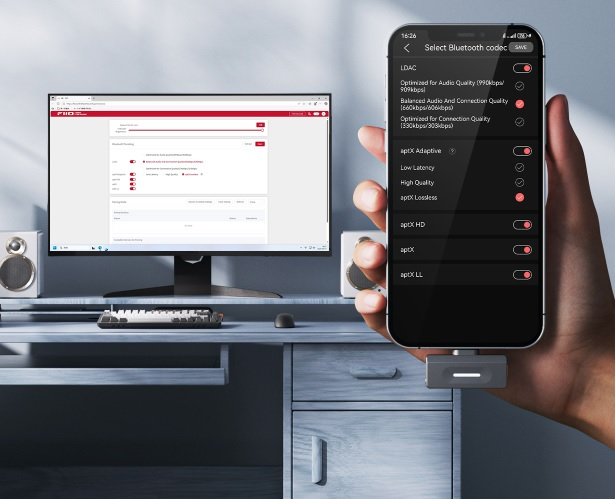 Ręka trzyma smartfon z ustawieniami kodeków Bluetooth, w tle biurko z monitorem.