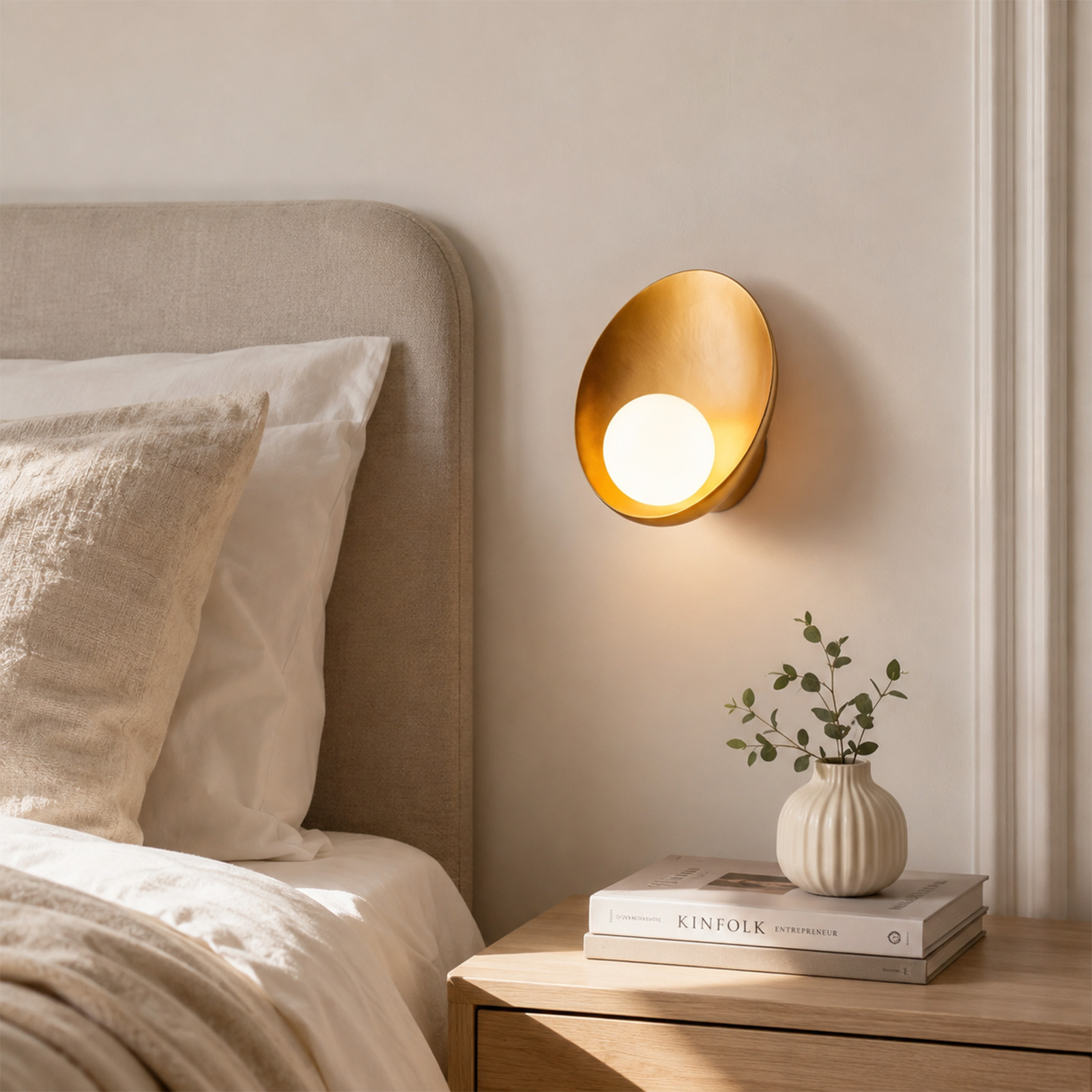 Cabecero de cama beige con aplique de pared dorado, mesita de noche, libros y un jarrón pequeño.
