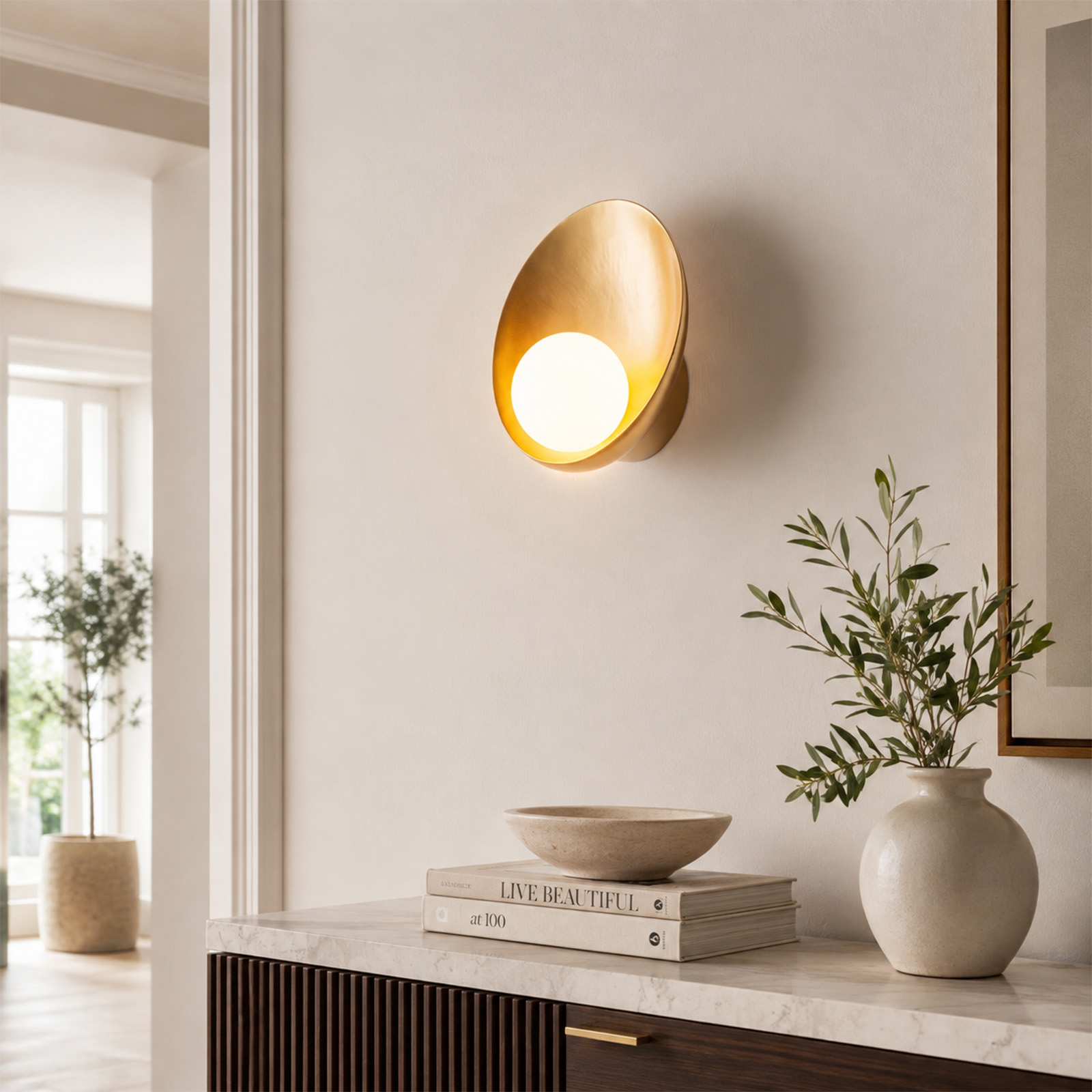 Un aplique de pared dorado sobre una mesa consola con libros, un cuenco y un florero.