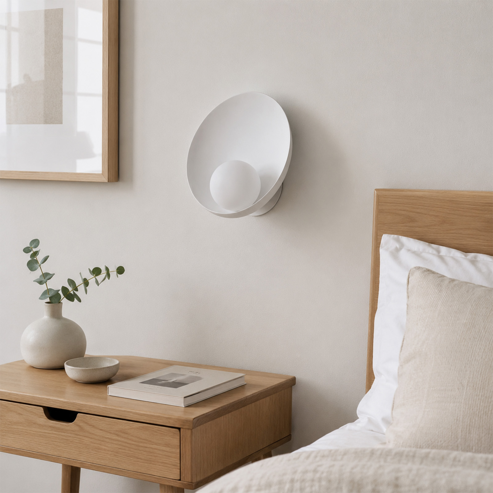 Un dormitorio minimalista con mesita de noche de madera, jarrón blanco, libro y una lámpara de pared moderna.