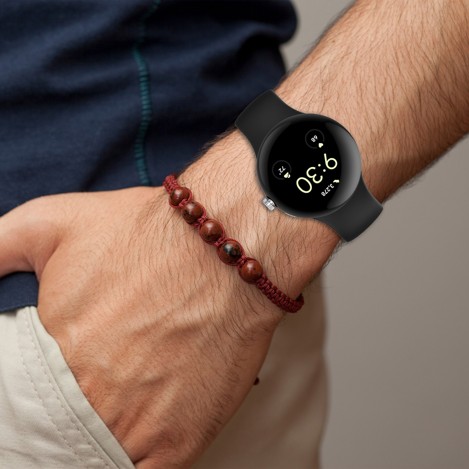 Arm mit einer schwarzen Smartwatch und einem braunen Perlenarmband, Hand in der Hosentasche.