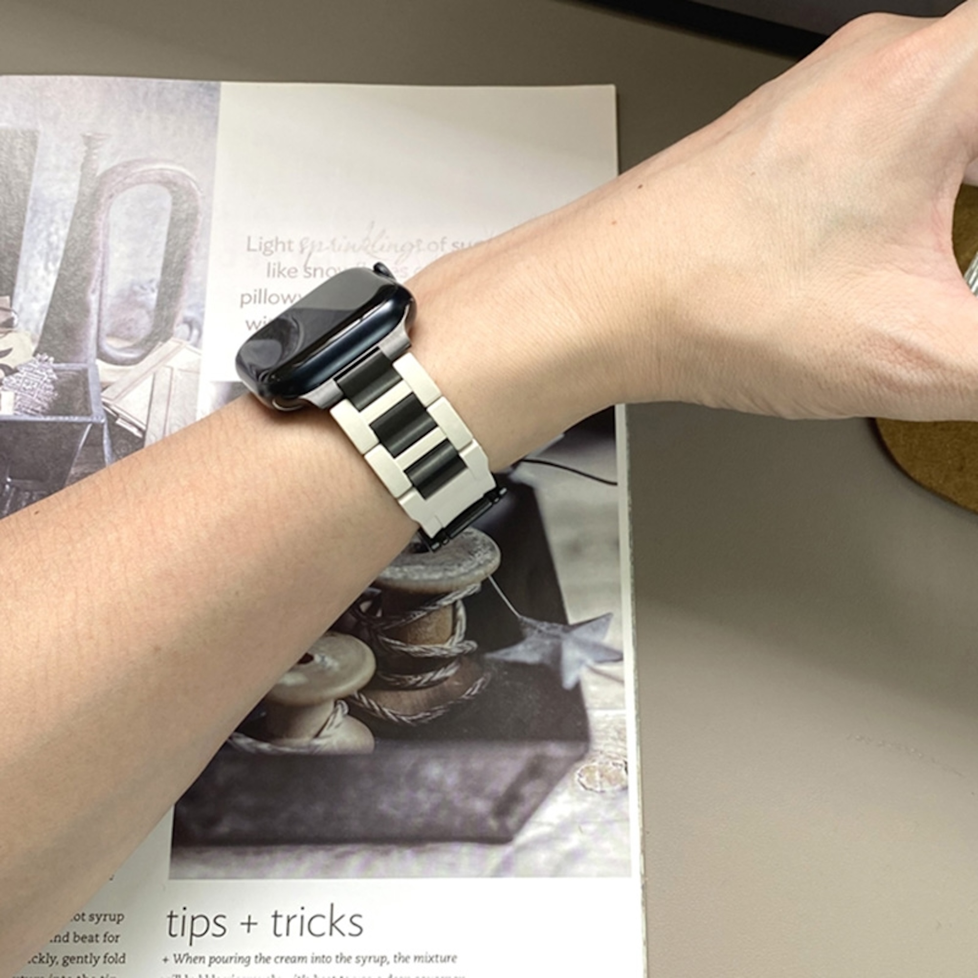 Eine Hand trägt eine Smartwatch mit schwarzem Gehäuse und einem weiß-schwarzen Gliederarmband über einer Zeitschrift.