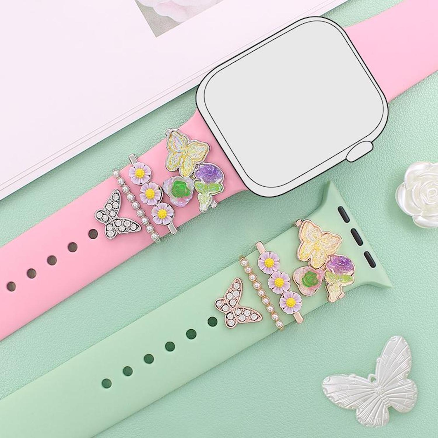 Rosa und grüne Uhrenarmbänder verziert mit Schmetterlings- und Blumen-Charms auf grünem Hintergrund.