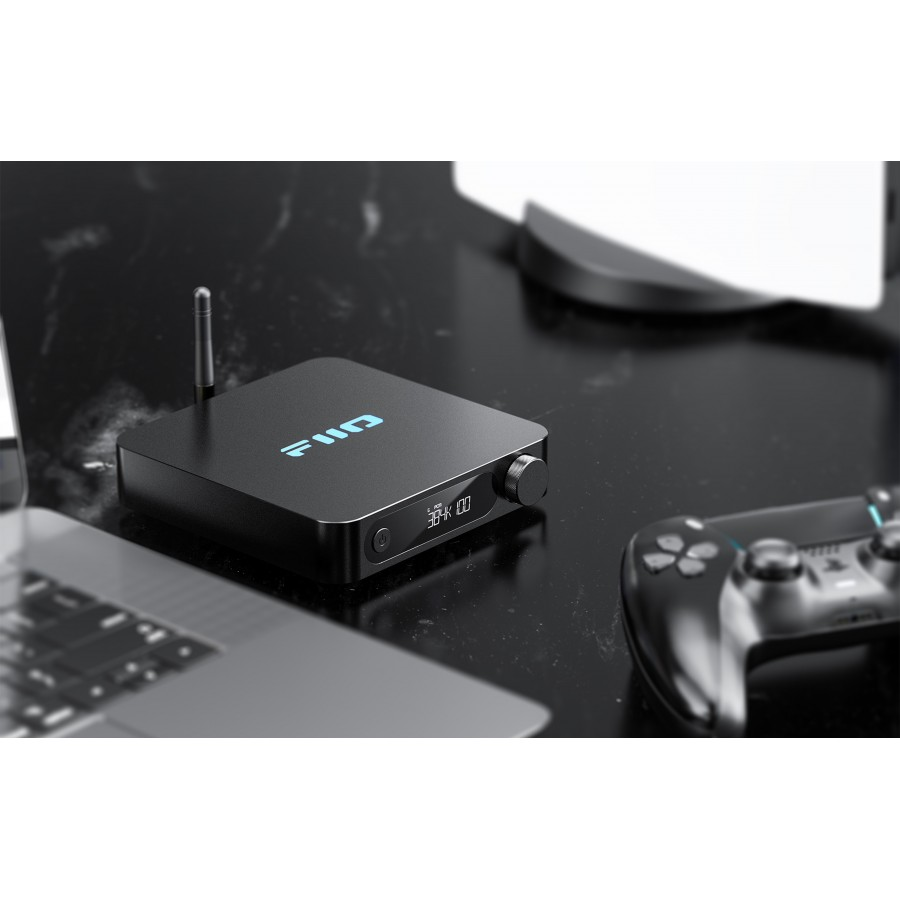 Un dispositivo de audio negro con logotipo azul y pantalla digital en un escritorio brillante junto a un mando y portátil.