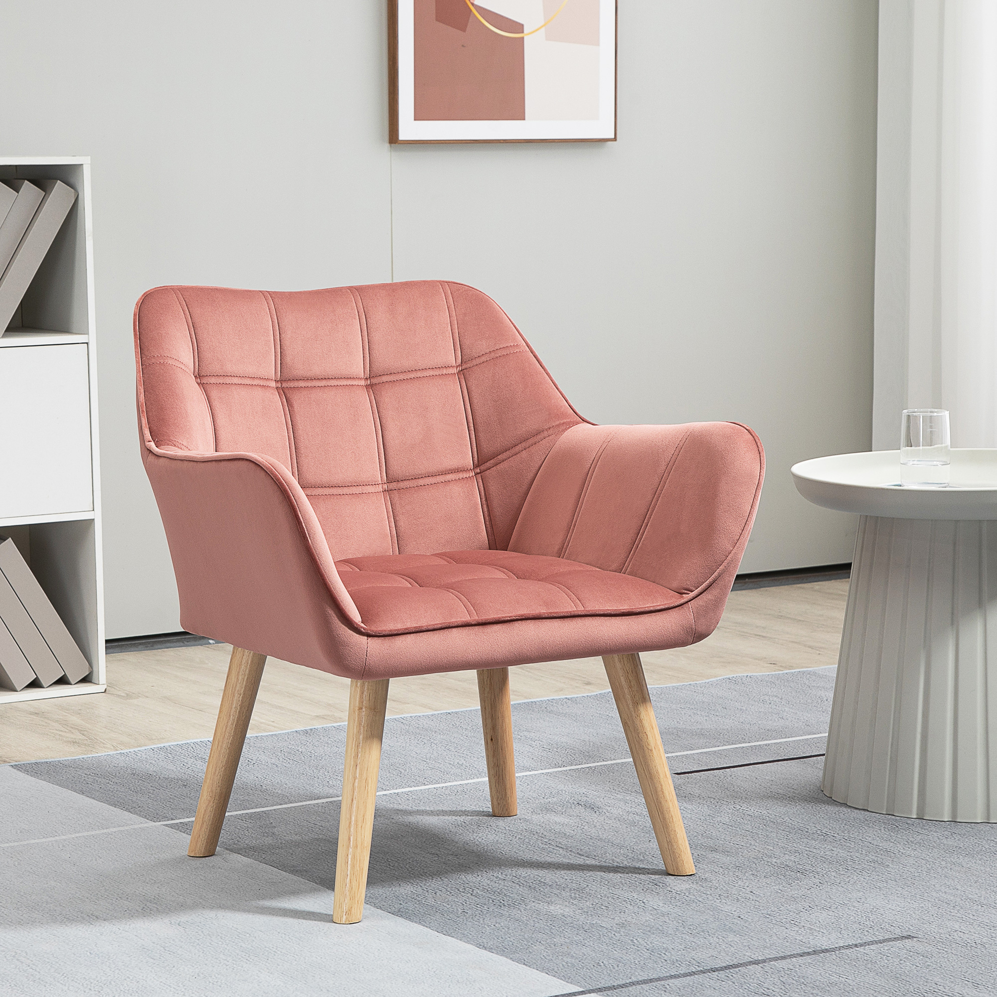 Una poltrona rosa trapuntata con gambe in legno su un tappeto grigio accanto a un tavolino bianco.