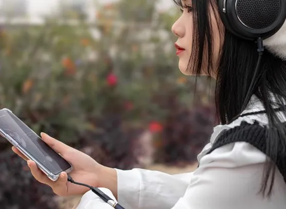 Una mujer de cabello negro usa grandes auriculares y sostiene un teléfono inteligente al aire libre.