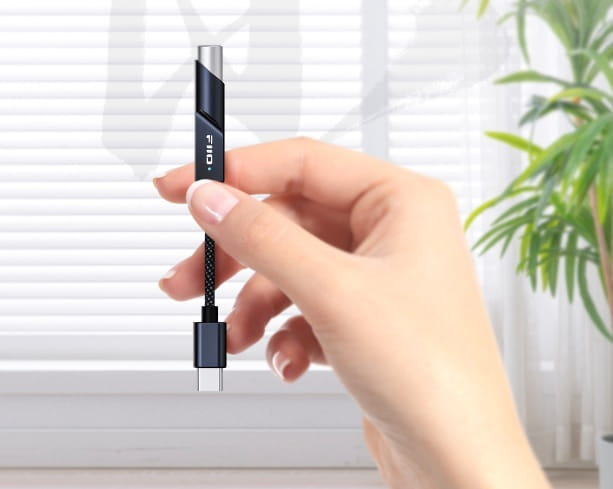 Una mano sostiene un adaptador USB-C frente a una ventana luminosa con plantas.