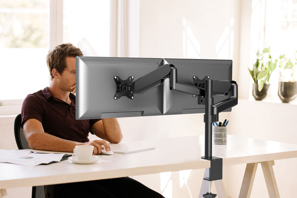 Een man zit aan een helder bureau voor twee monitoren op een verstelbare arm.
