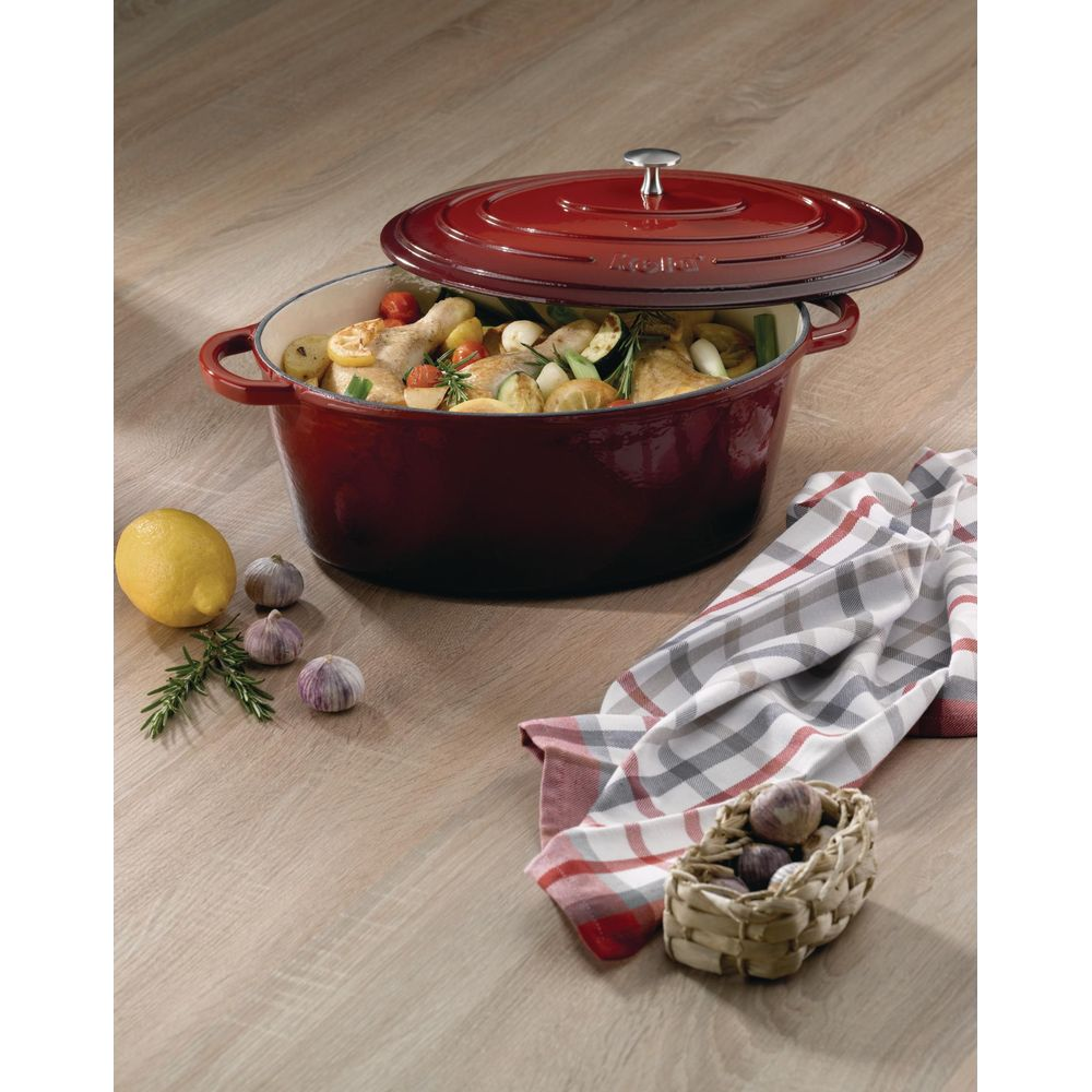 Ein roter Gusseisentopf mit Hähnchen und Gemüse steht auf einem Holztisch, neben Zitrone, Knoblauch und einem karierten Tuch.