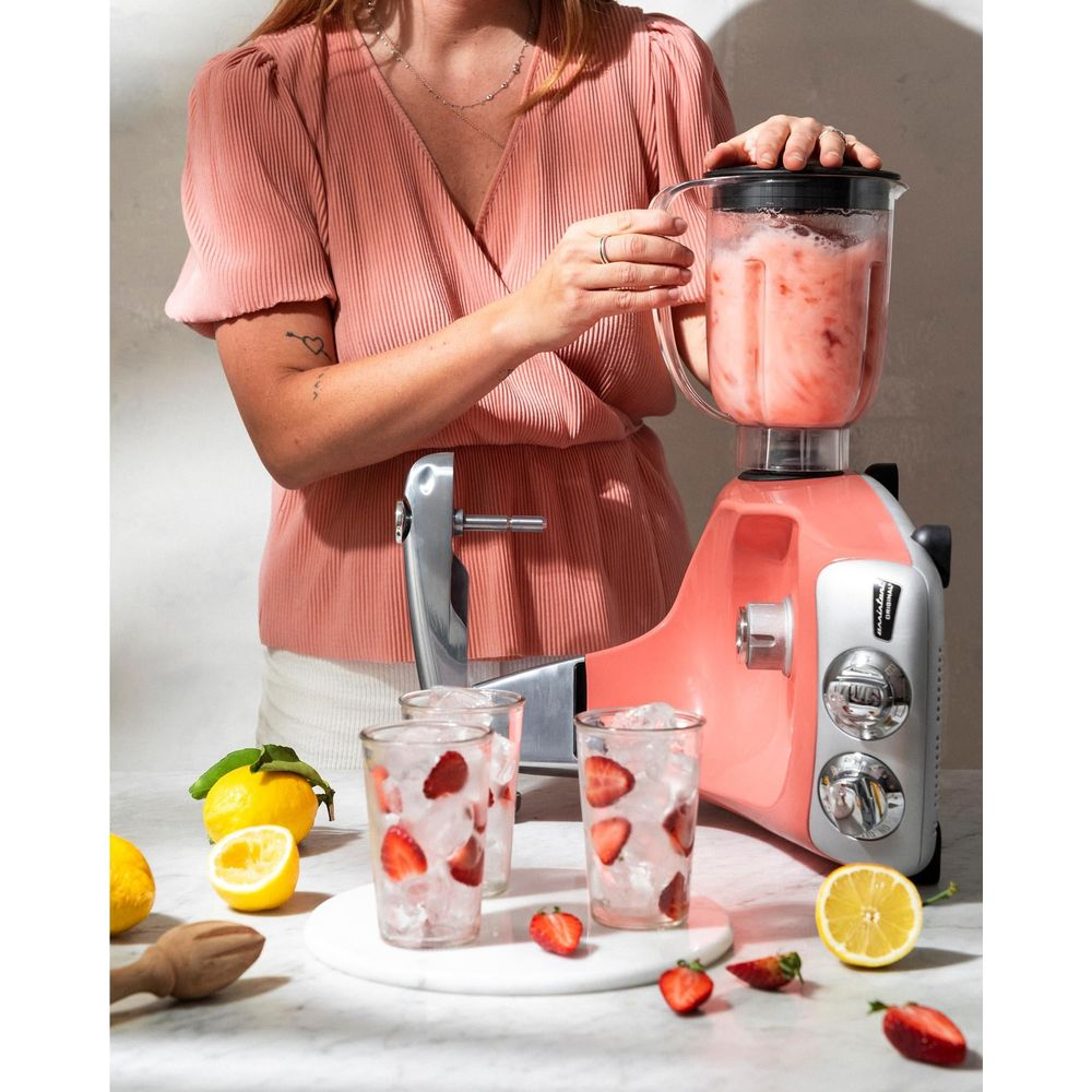 Eine Frau in einem rosa Oberteil bereitet einen rosafarbenen Smoothie in einem Mixer zu, umgeben von Zitronen und Erdbeeren.