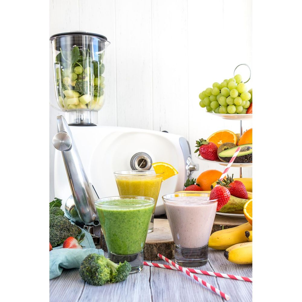 Drei Smoothies in Gläsern, ein weißer Mixer, frisches Obst und Gemüse stehen auf einem Holztisch.