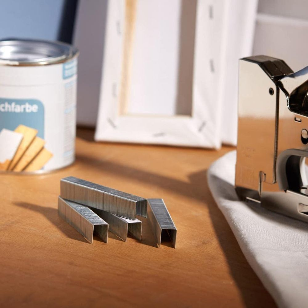 Metallklammern liegen auf einer Holzoberfläche neben einem Tacker, einer Farbdose und Leinwänden.
