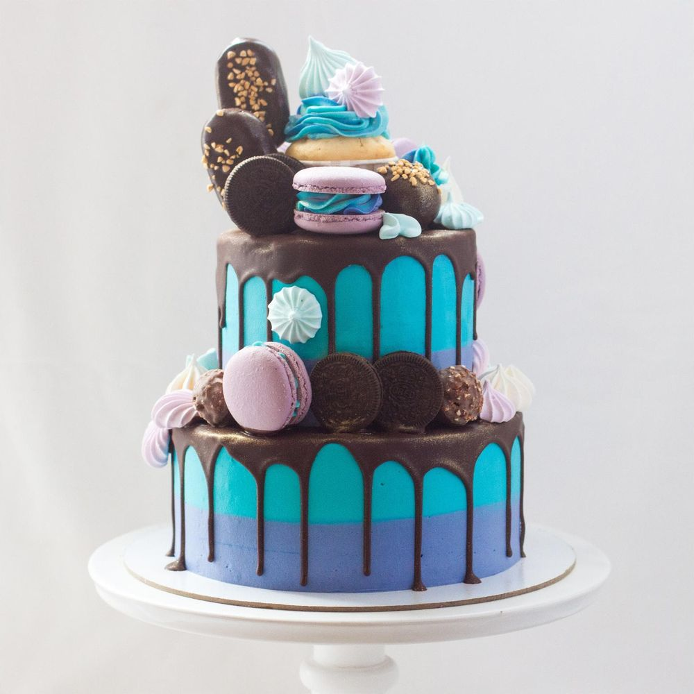 Zweistöckige Torte in Blau und Lila mit Schokoladenglasur, gekrönt von Macarons, Keksen und Baiser.