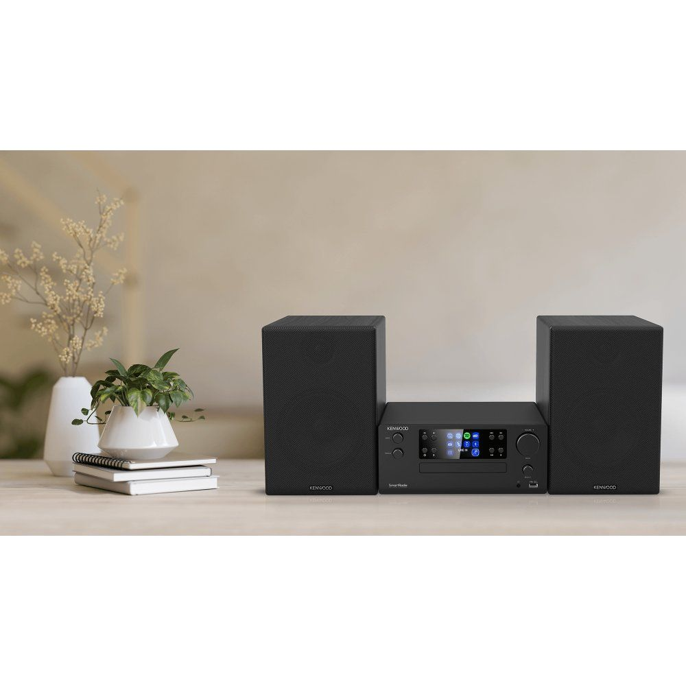 Sistema hi-fi Kenwood nero con due altoparlanti su un tavolo, accanto a piante in vaso e libri.