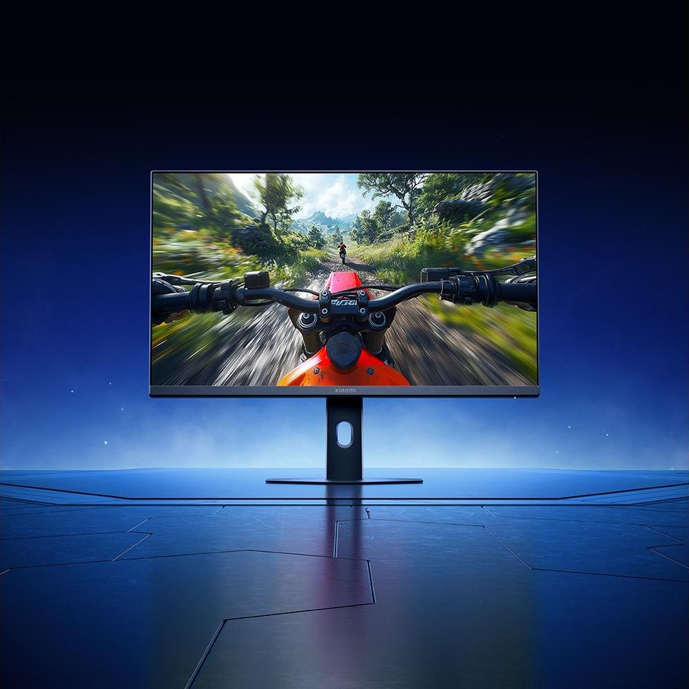 Un monitor Xiaomi en una mesa azul muestra un juego de carreras de motos con vista del manillar en una pista verde.
