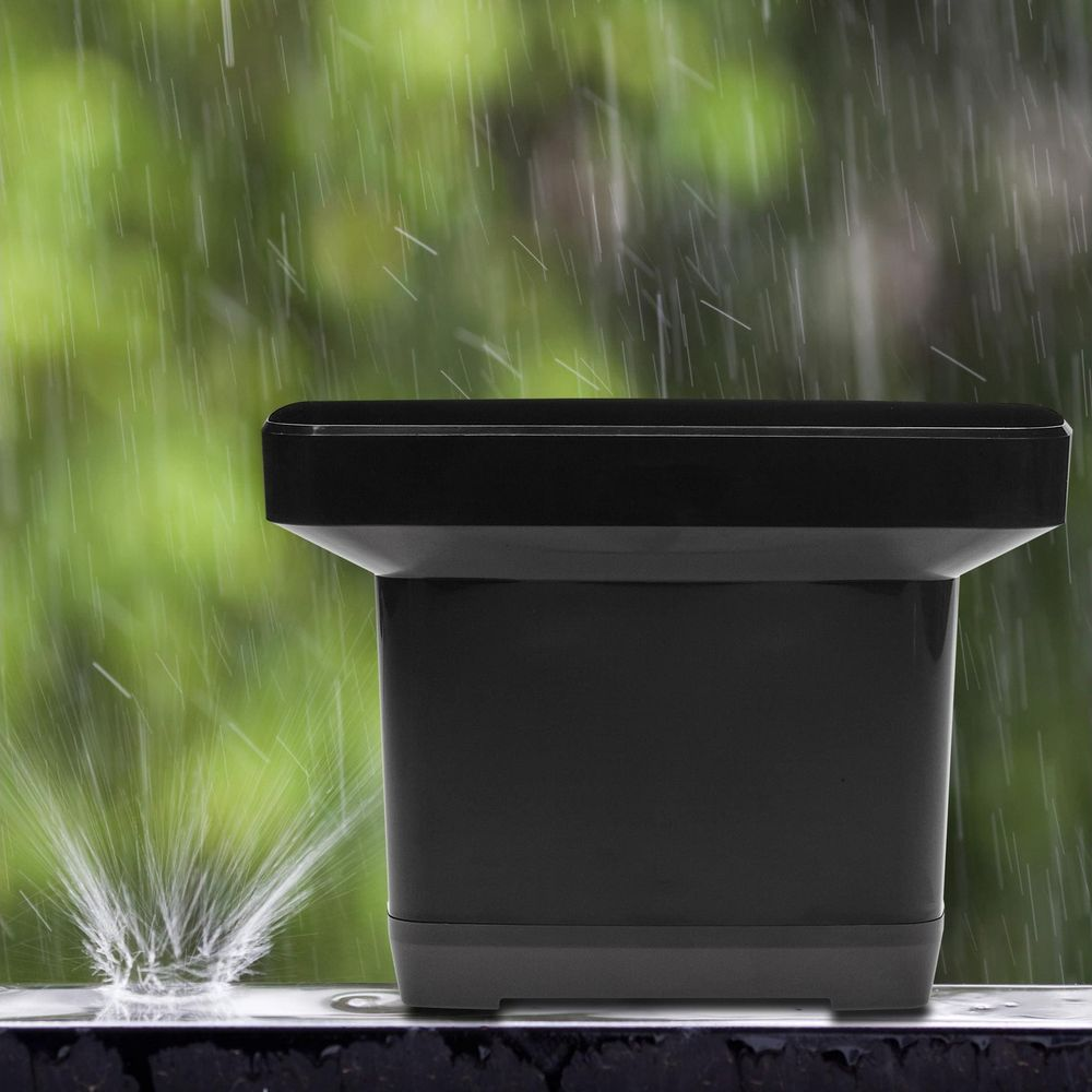 Ein schwarzer Regensensor steht im starken Regen vor einer grünen Naturkulisse.