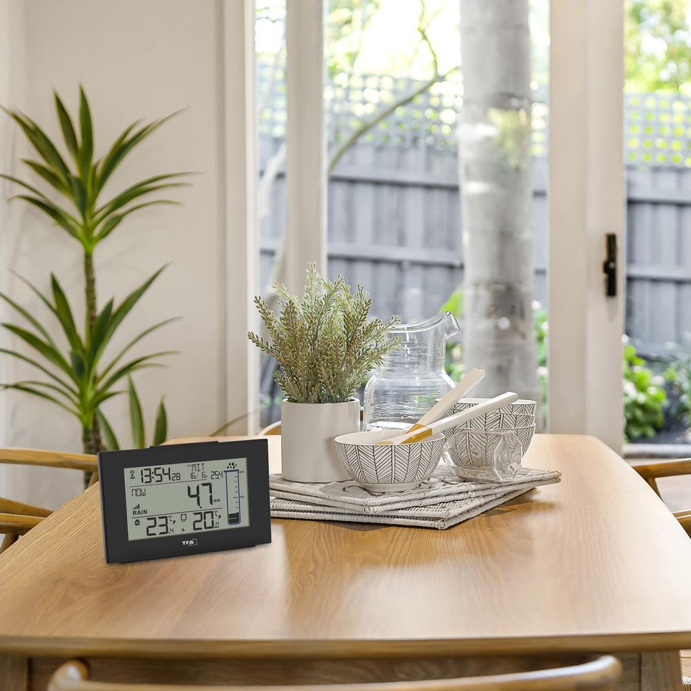 Wetterstation auf Holztisch in hellem Esszimmer mit Pflanzen und Schüsseln im Hintergrund.