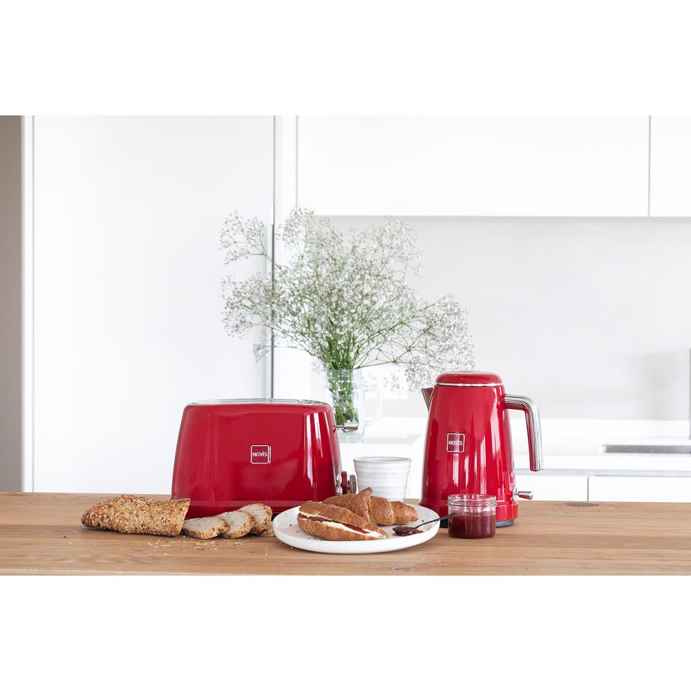 Roter Toaster und Wasserkocher auf Holztisch mit Brot, Croissants und Blumen im Hintergrund.