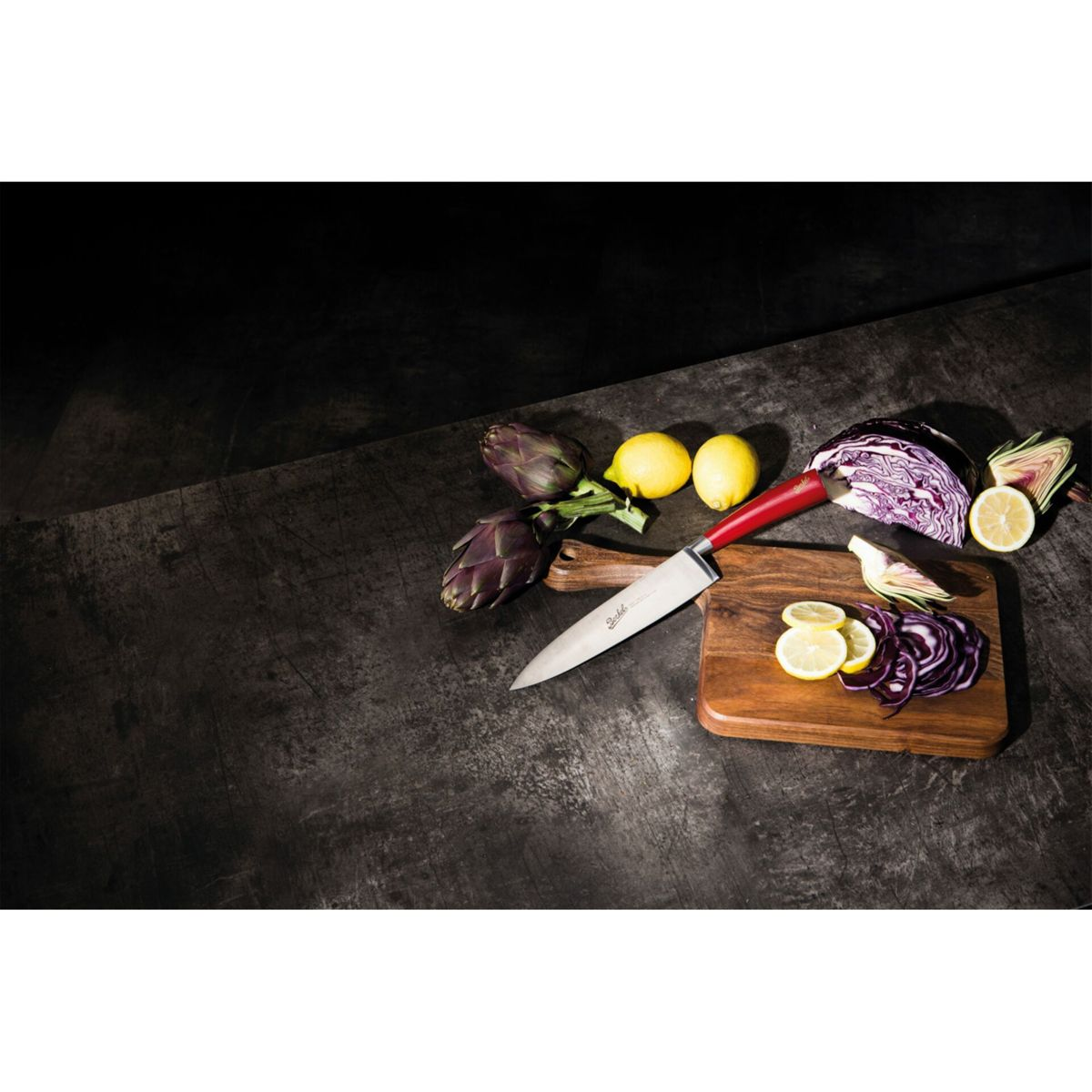 Coltello con manico rosso su tagliere con limoni, cavolo rosso e carciofi su superficie scura.