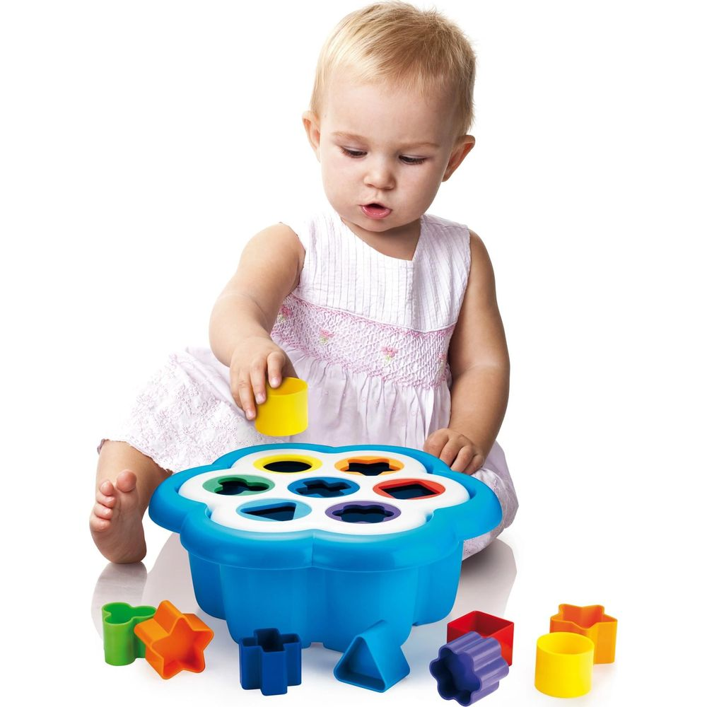 Kleinkind spielt mit blauem Formensortierer und bunten Klötzen auf weißem Grund.