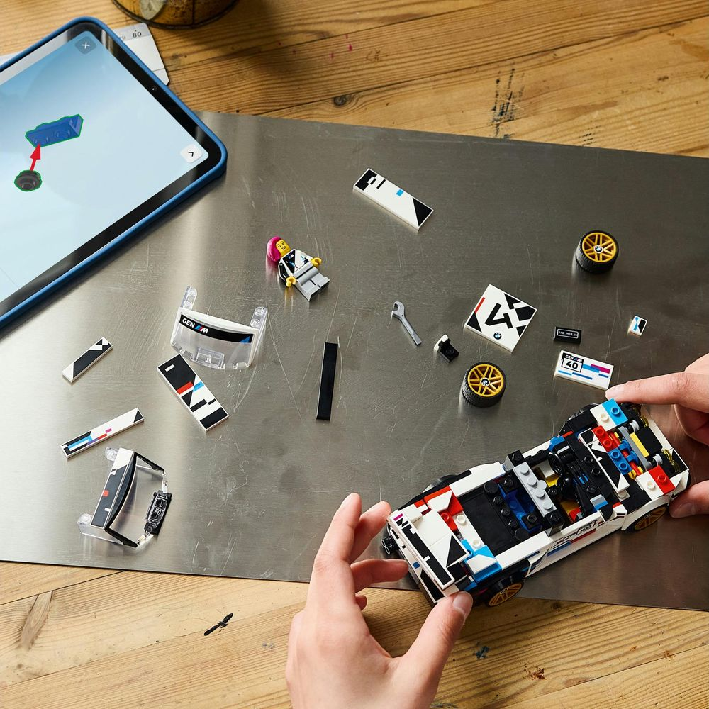 Le mani assemblano un'auto Lego su una superficie metallica, circondate da mattoncini, una minifigura e un tablet.