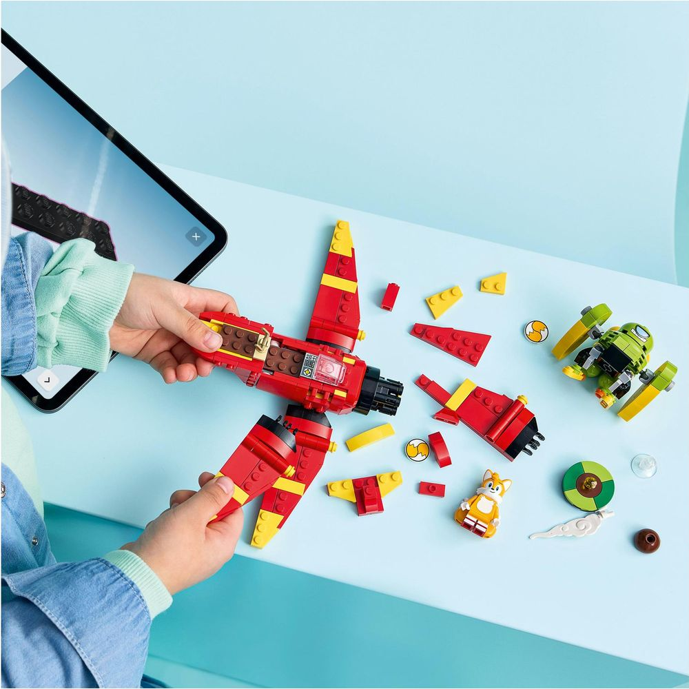 Un niño construye un avión rojo de LEGO junto a una tableta, piezas sueltas, un robot verde y una figura de Tails.