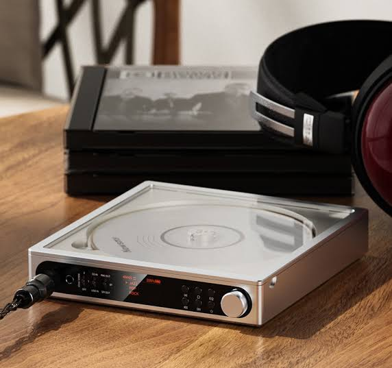 Un reproductor de CD plateado y auriculares sobre una mesa de madera, con cajas de CD detrás.