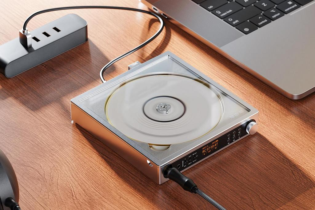 Un reproductor de CD plateado con cubierta transparente y cable se encuentra junto a una laptop en un escritorio de madera.