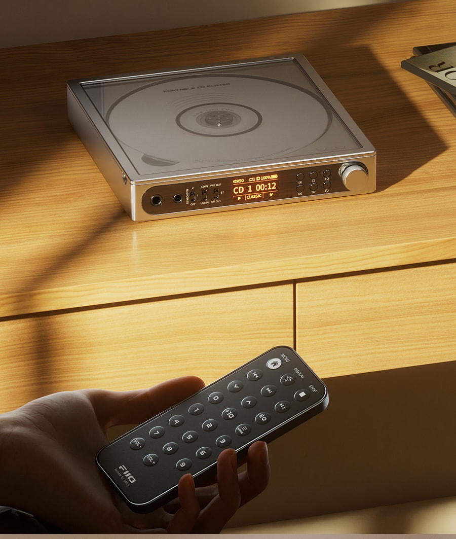 Una mano sostiene un control remoto frente a un reproductor de CD portátil plateado en una mesa de madera.