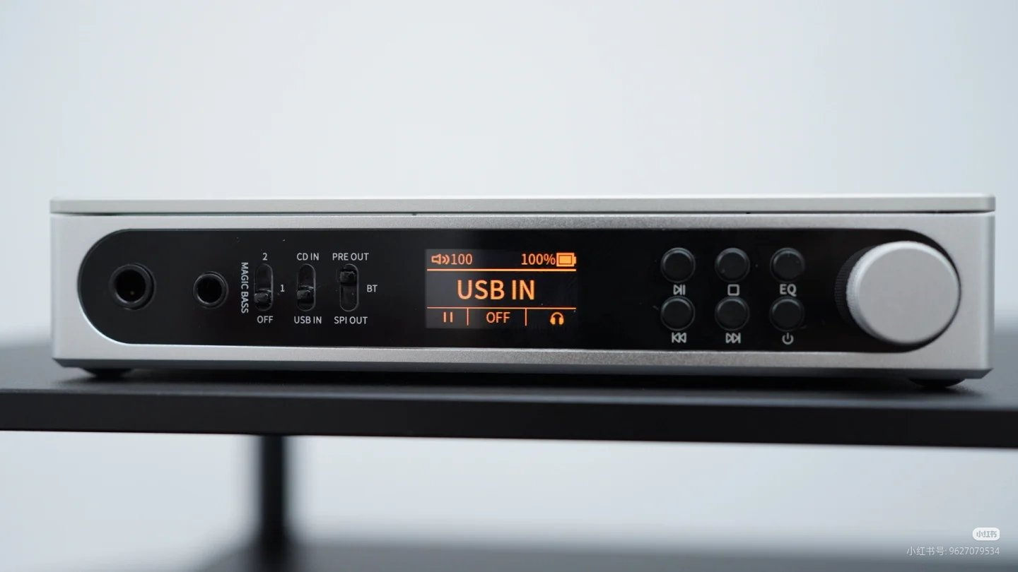Srebrne urządzenie audio z czarnym panelem, pomarańczowym wyświetlaczem, przyciskami i pokrętłem.