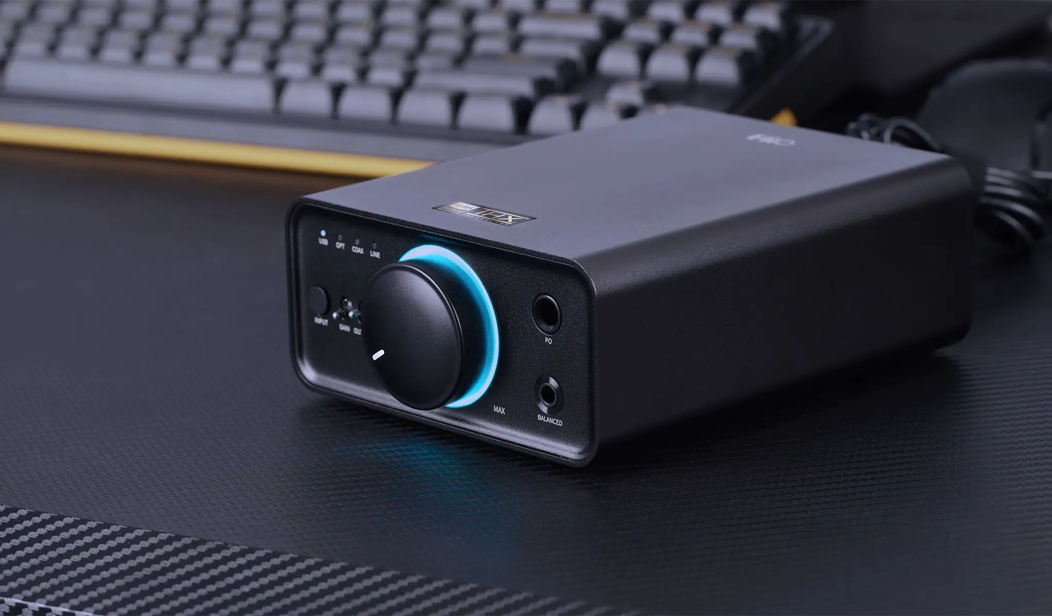 Un amplificador de audio negro con una perilla azul brillante se encuentra en un escritorio frente a un teclado.